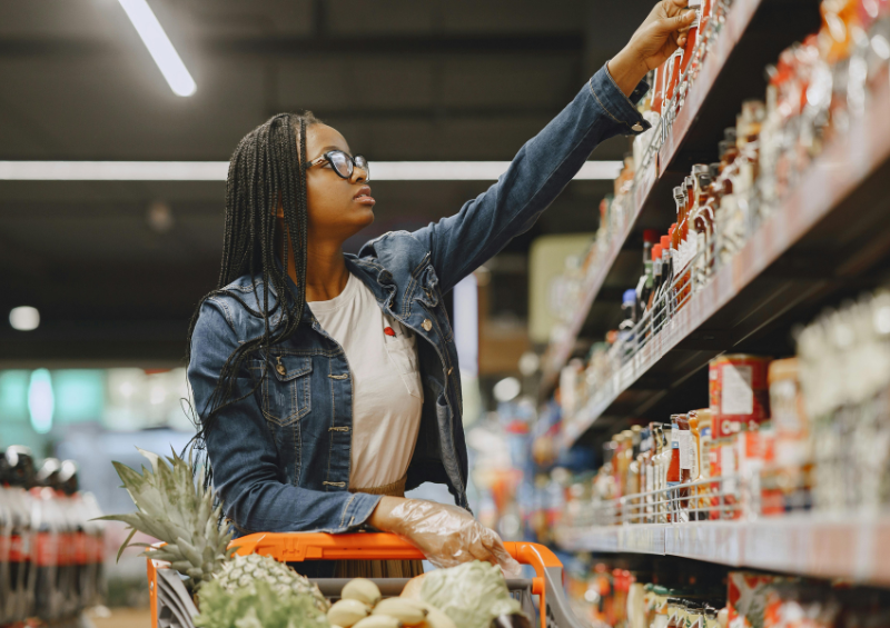 mulher negra com tranças e óculos escolhendo produtos no mercado e com um carrinho de compras cheio