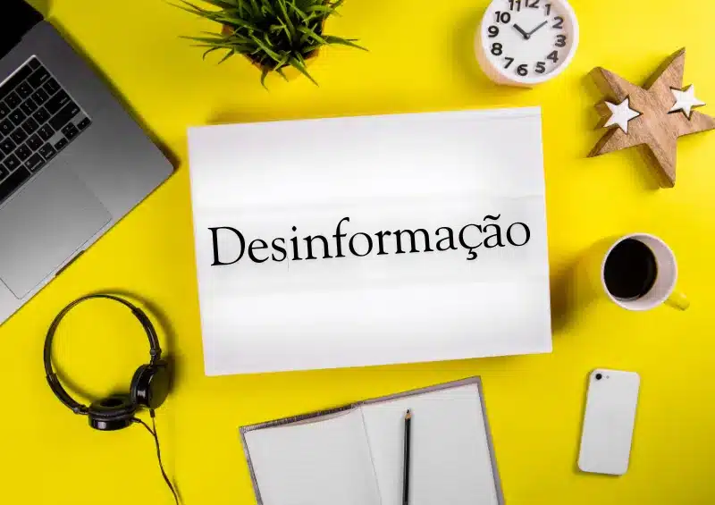 fundo amareço com xícara de café, fones de ouvido, estrela do mar e relógio. No meio tem uma folha branca escrito desinformação,